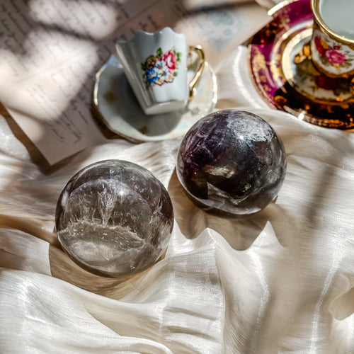 Two crystal balls of fluorite with mica spheres on a textured surface with a floral teacup in the background.